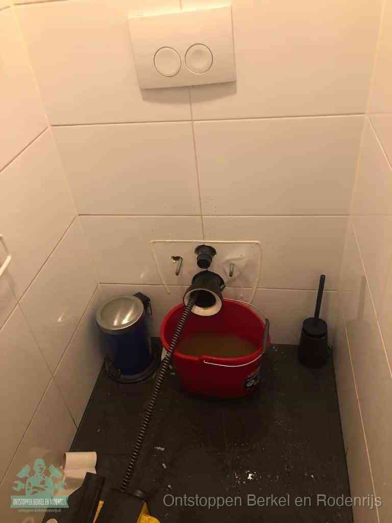 Een ontstopte wand-wc met verwijderde toiletpot waar een ontstoppingsveer in de wanduitgang verdwijnt, het achterhalen waar de verstopping zit in Berkel en Rodenrijs