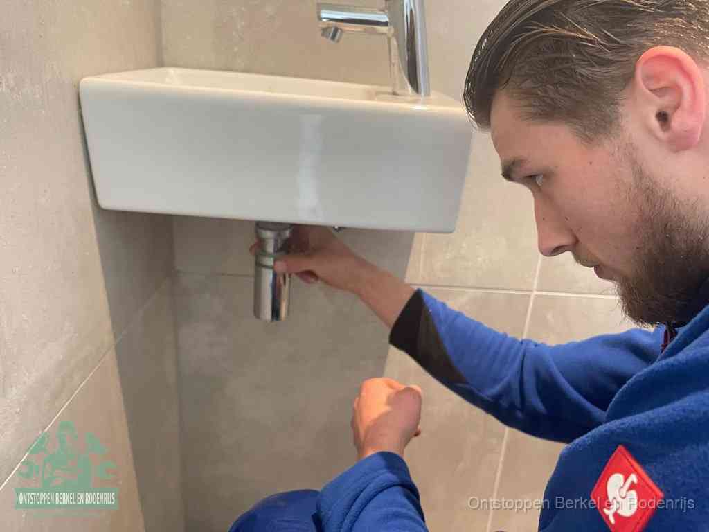 Een monteur draait met de hand de zwanenhalssifon onder een fonteinwasbak in de toiletruimte los om de bron van rioollucht in Berkel en Rodenrijs te vinden