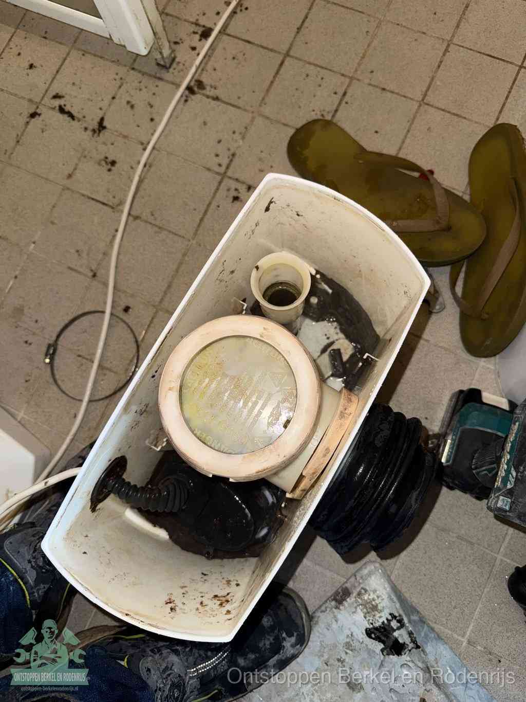 Een gedemonteerde toiletpot op de vloer met ontstoppingsveer en gereedschap ernaast, het verwijderen van een vastzittend voorwerp uit de wc in Berkel en Rodenrijs