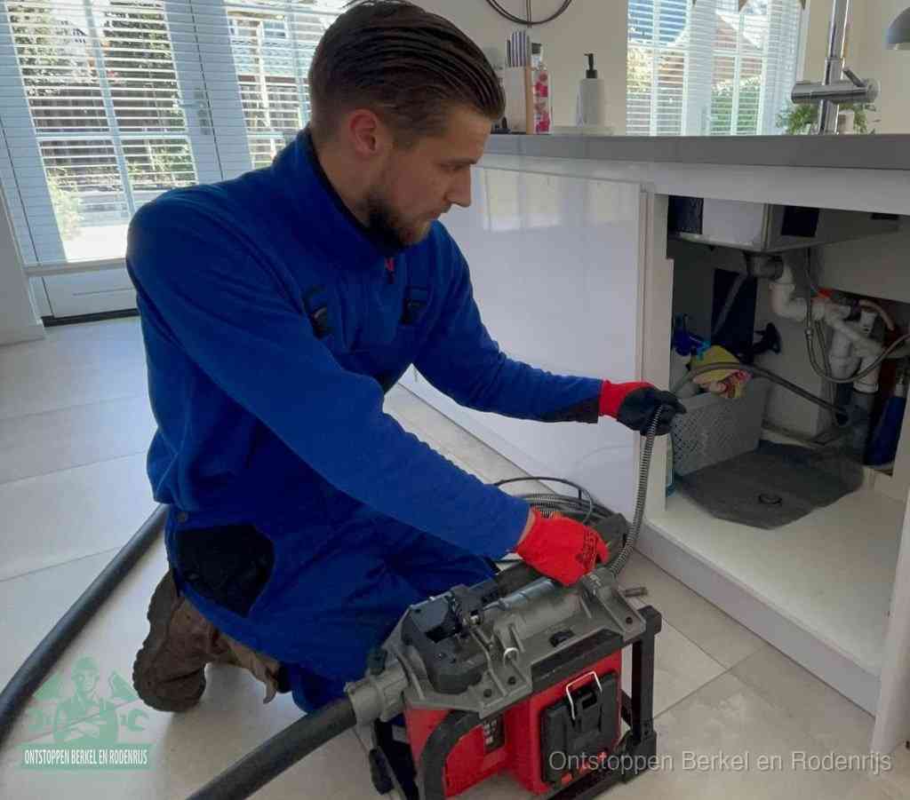 Een monteur met rode handschoenen knielt in een keuken en voert een elektrische ontstoppingsveer in tijdens het professioneel afvoer ontstoppen in Berkel en Rodenrijs
