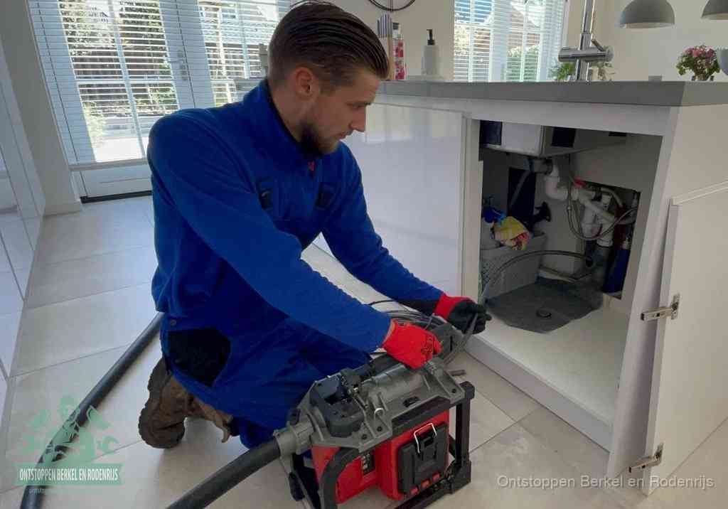 Een monteur voert een professionele elektrische ontstoppingsmachine in de afvoer onder de spoelbak tijdens het gootsteen ontstoppen in Berkel en Rodenrijs